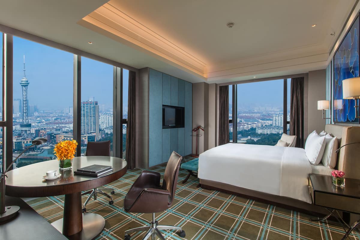 Une chambre d’hôtel avec un grand lit, un bureau avec une chaise, une petite table ornée d’un vase de fleurs et une vue sur le paysage urbain.