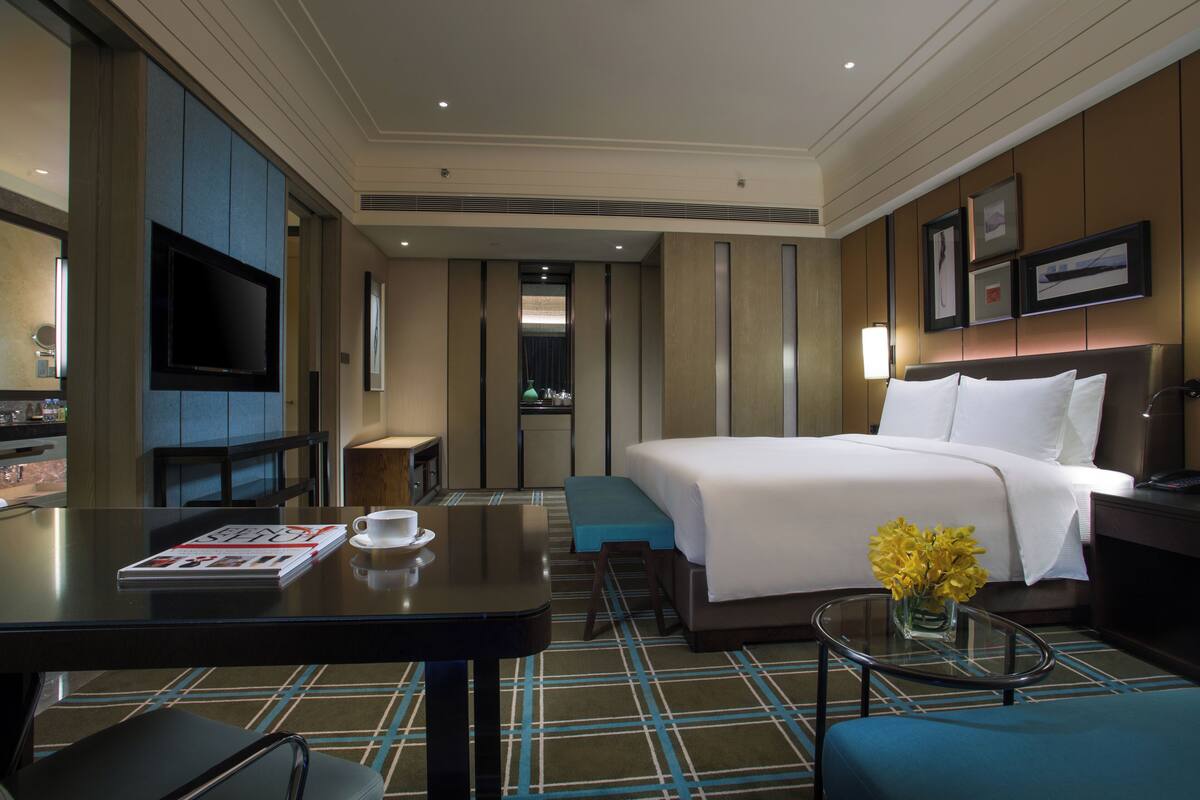 Une chambre d’hôtel avec un lit, une télévision, un bureau avec un magazine et une tasse, et une petite table avec des fleurs.