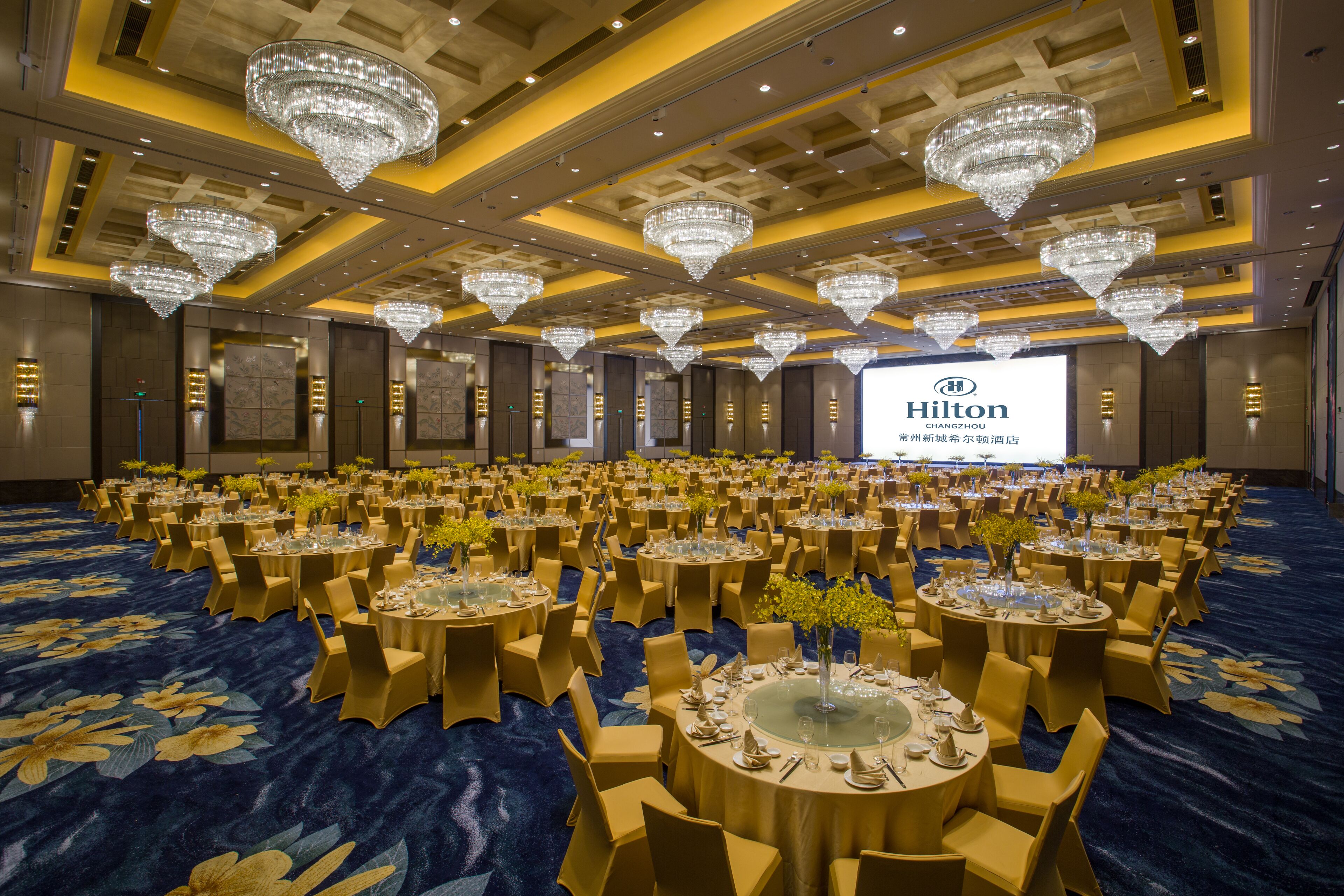 Une vaste salle de banquet avec des tables rondes, des chaises dorées et des lustres.