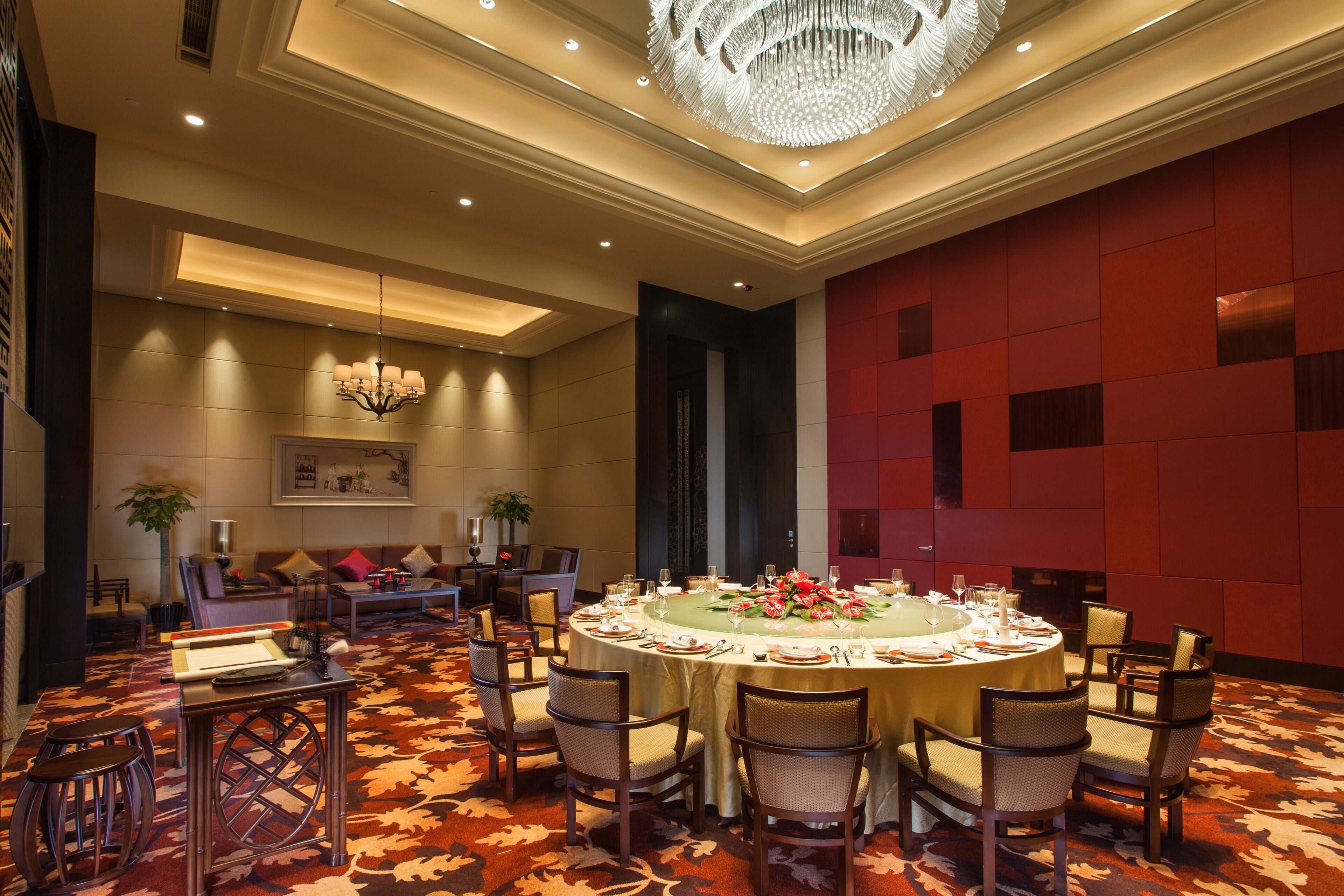 A hotel lobby with a round dining table set for six, surrounded by chairs, a chandelier, and a red accent wall.