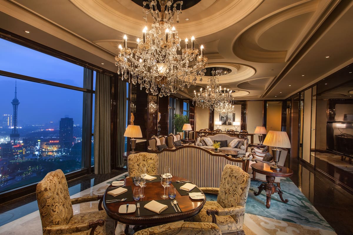 Une chambre d’hôtel luxueuse, dotée d’un grand lustre, d’une table à manger dressée pour quatre personnes et d’un canapé. Une grande fenêtre offre une vue sur la ville.