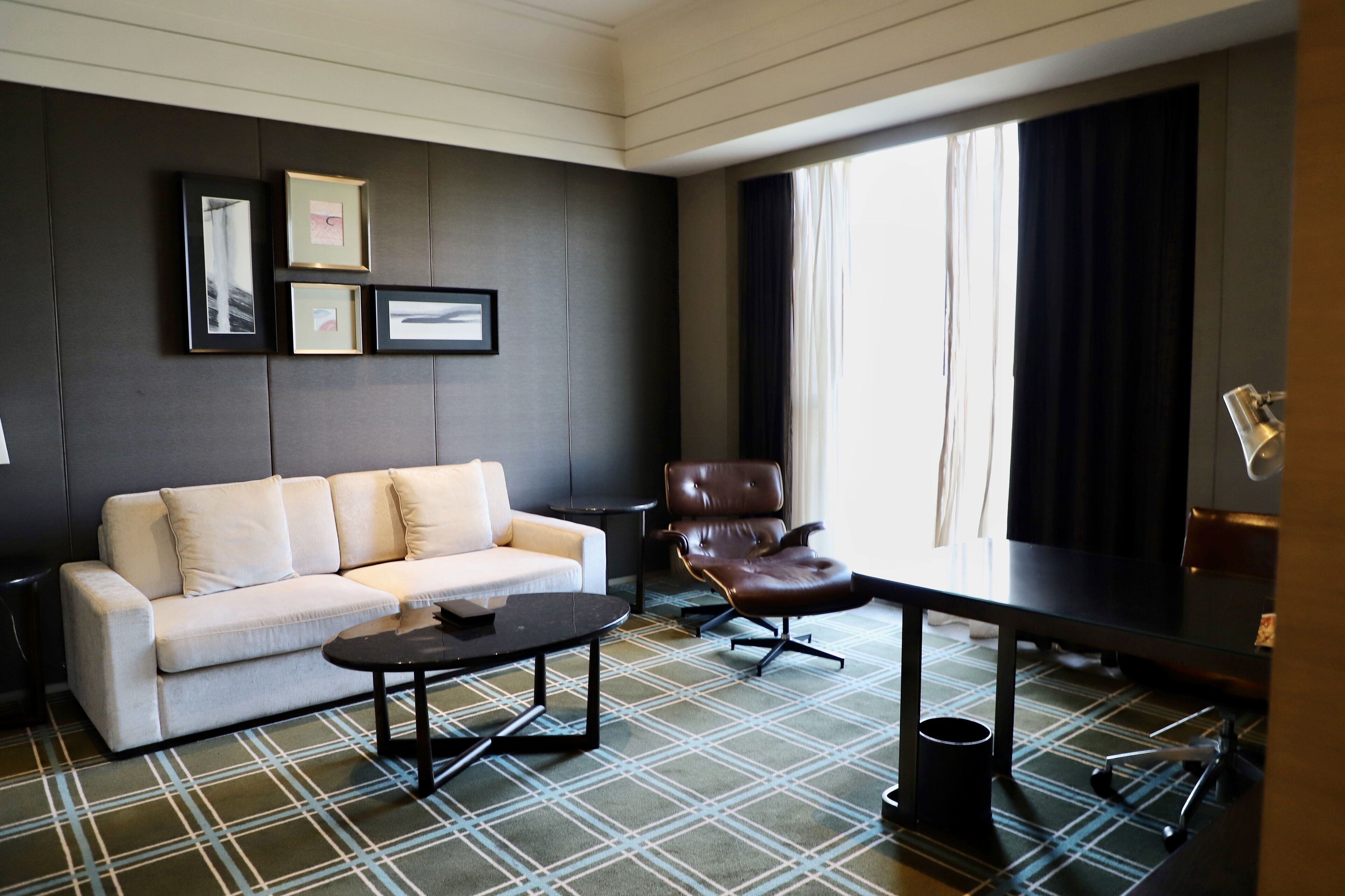 A modern living room with a sofa, a coffee table, a leather armchair, and a dining table. There are framed pictures on the wall and a window with curtains.