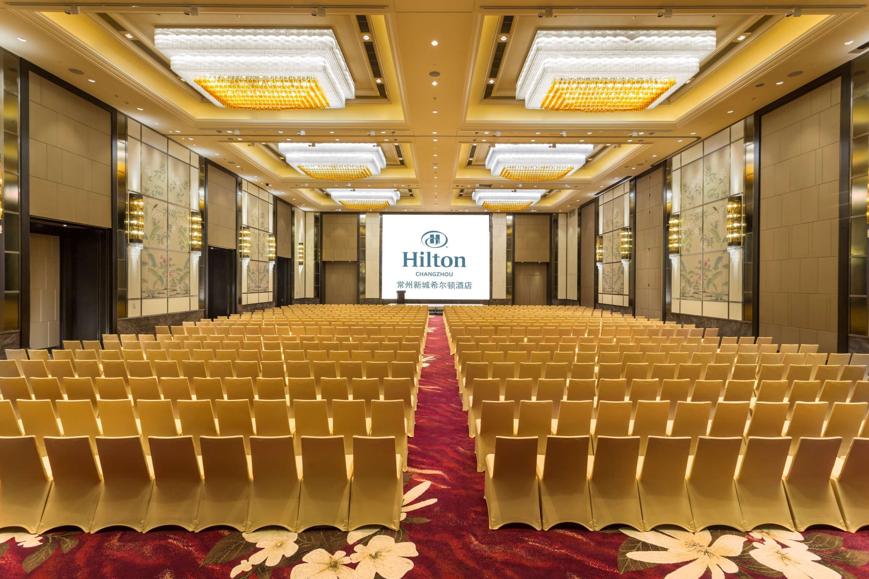 A large, well-lit conference room with rows of chairs and a red carpet. A Hilton logo is visible on the back wall.
