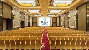 A large, well-lit conference room with rows of chairs and a red carpet. A Hilton logo is visible on the back wall.