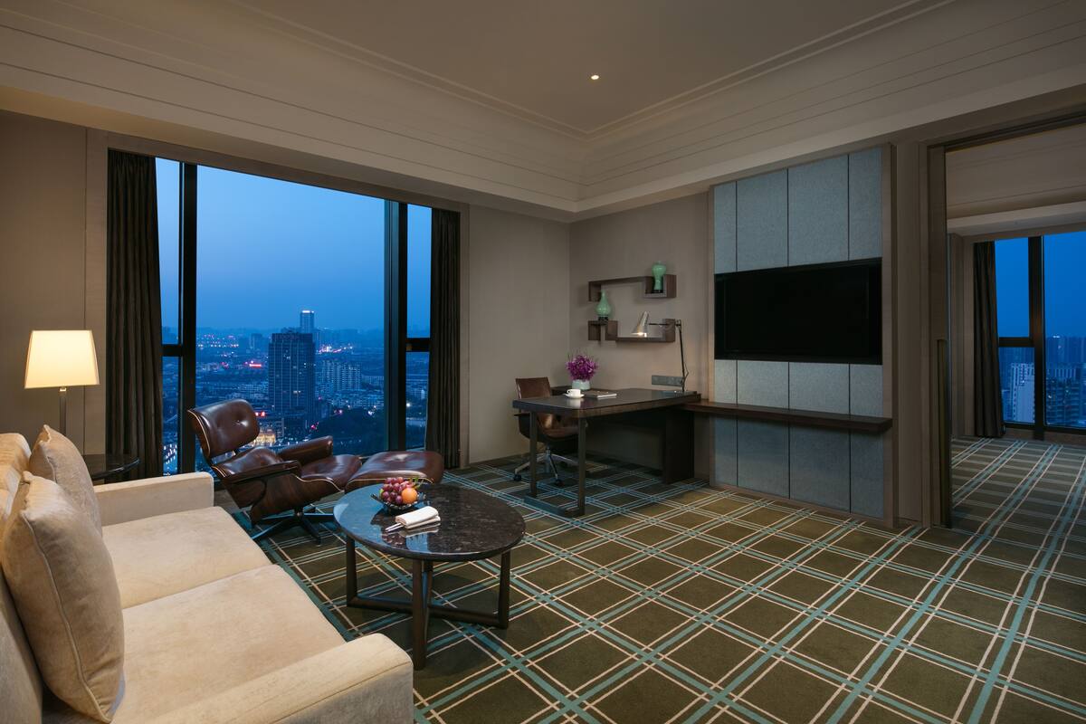 Une chambre d’hôtel dotée d’une grande fenêtre donnant sur la ville, d’un canapé, d’une table basse, d’un bureau et d’un téléviseur à écran plat.