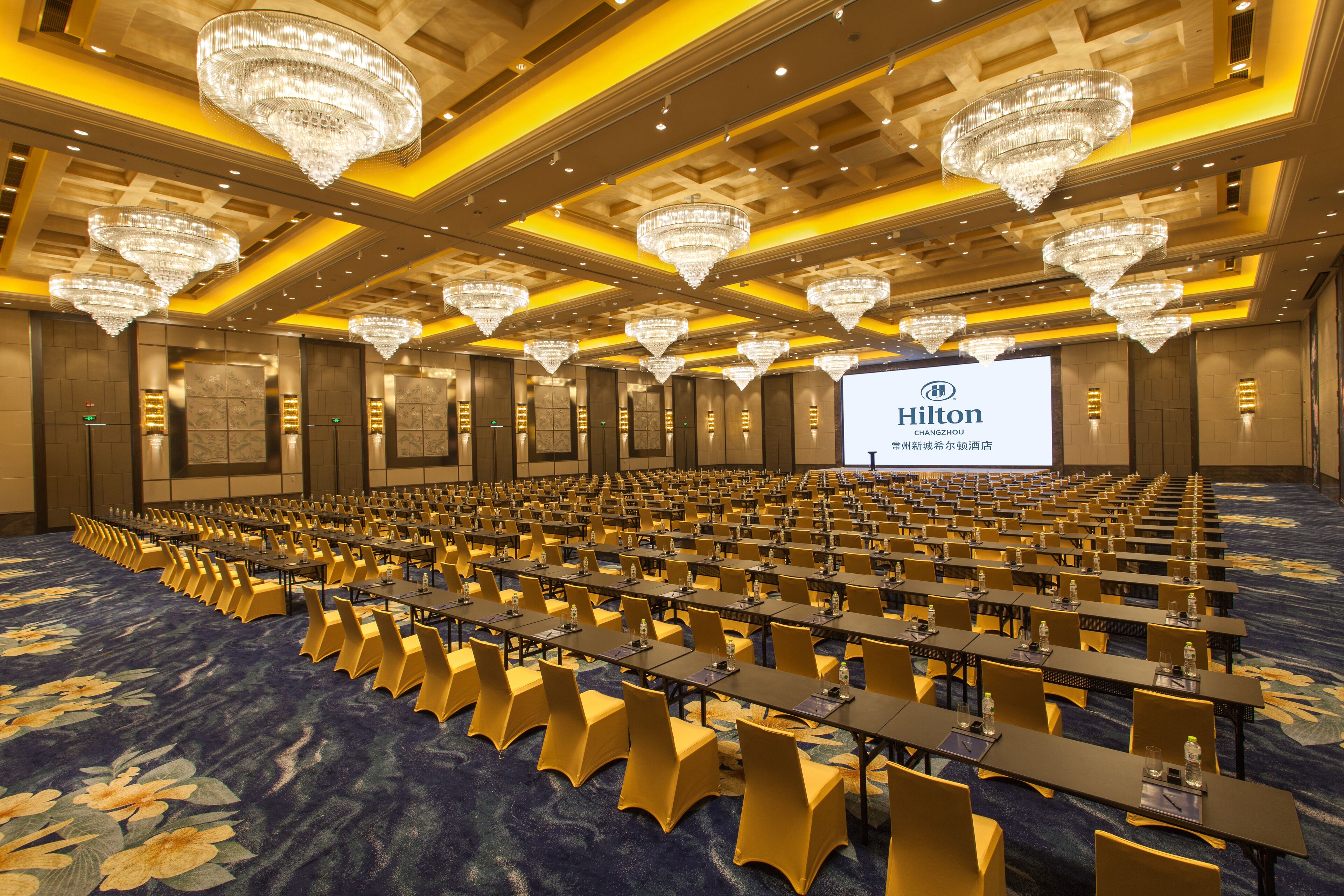 A large conference room with rows of tables and chairs, chandeliers, and a screen displaying 'Hilton CHANGZHOU'.