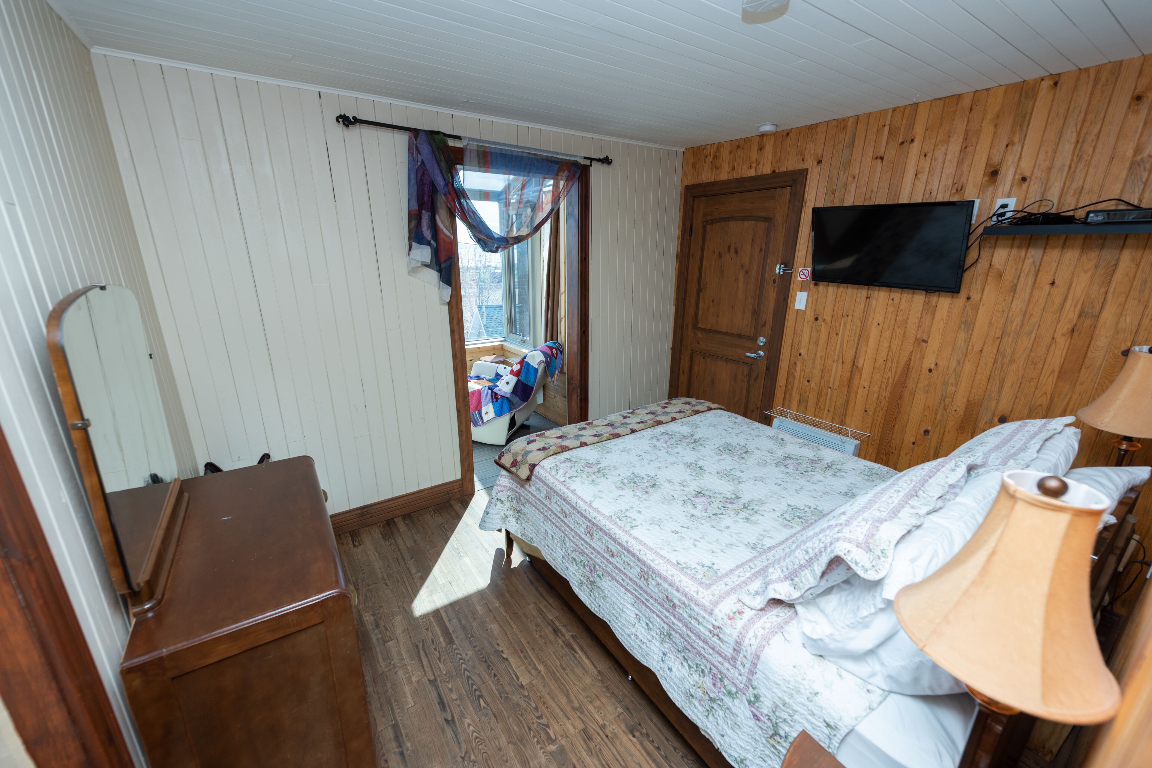 A bedroom with a wooden floor, a bed with a floral comforter, a wooden dresser, a television mounted on the wall, and a window with curtains.