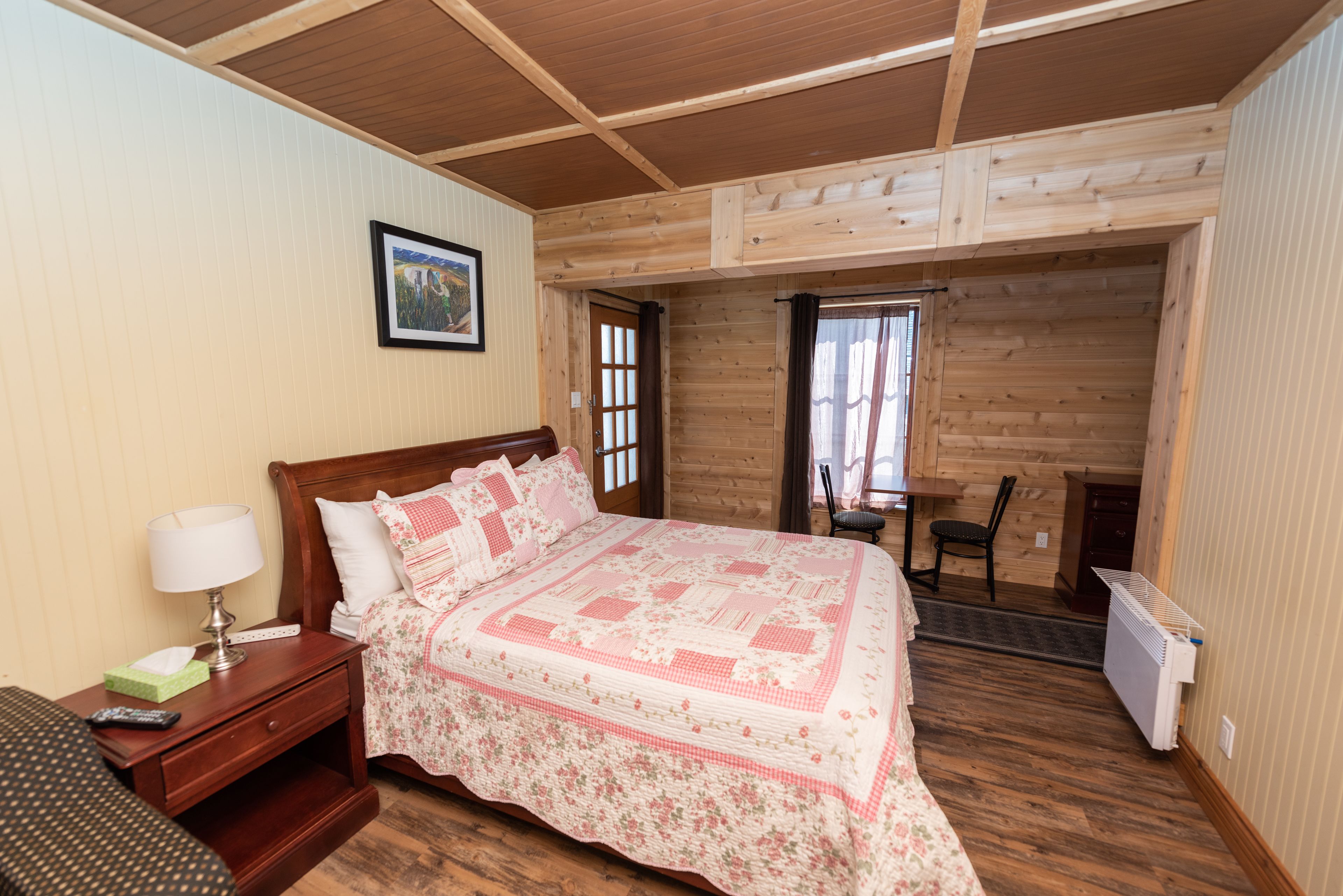 A bedroom with a wooden ceiling, a bed with a floral quilt, a bedside table with a lamp, and a chair near the window.