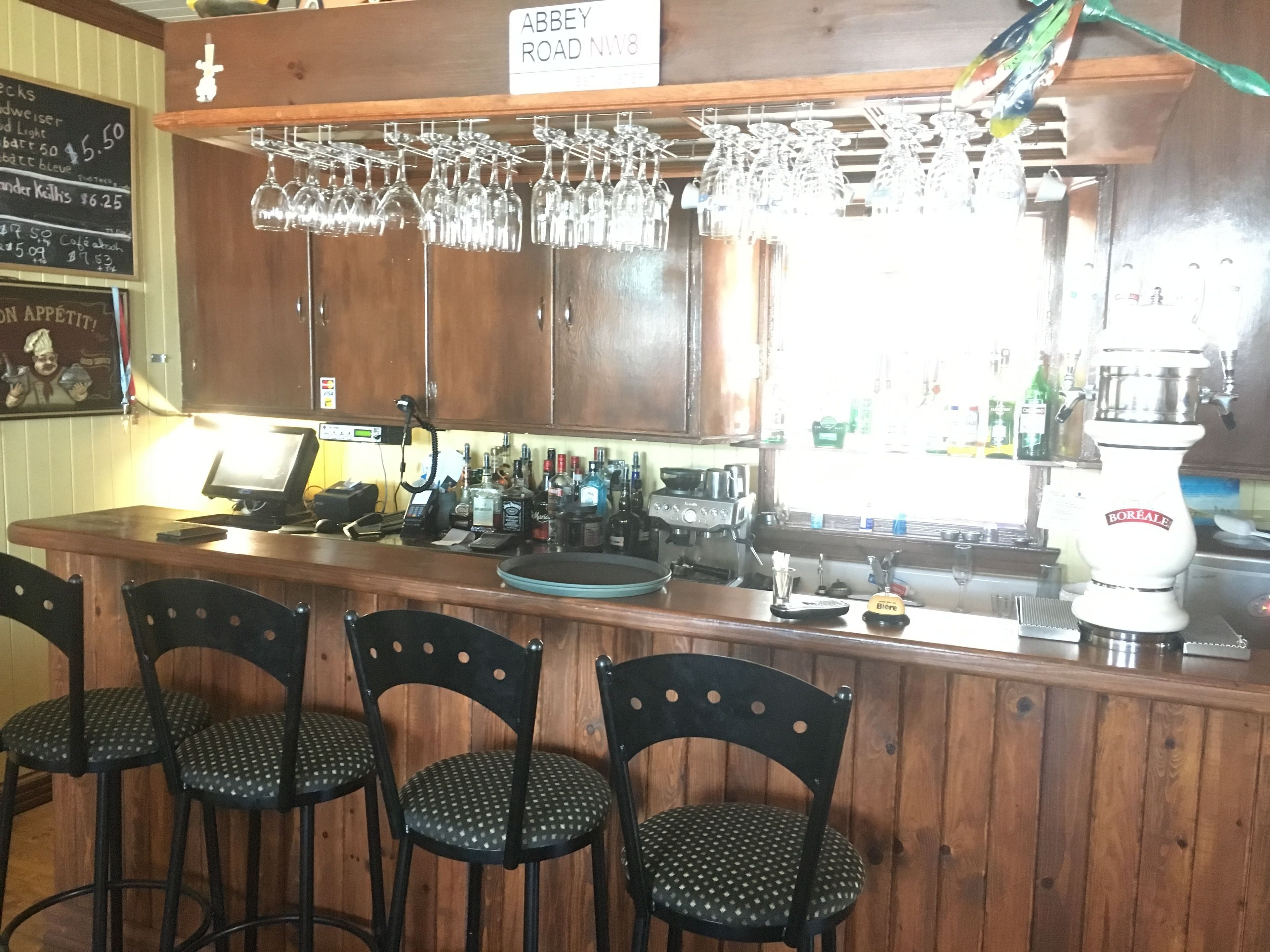 Un espace bar avec des meubles en bois, des verres à vin suspendus, une caisse enregistreuse et une tireuse à bière.