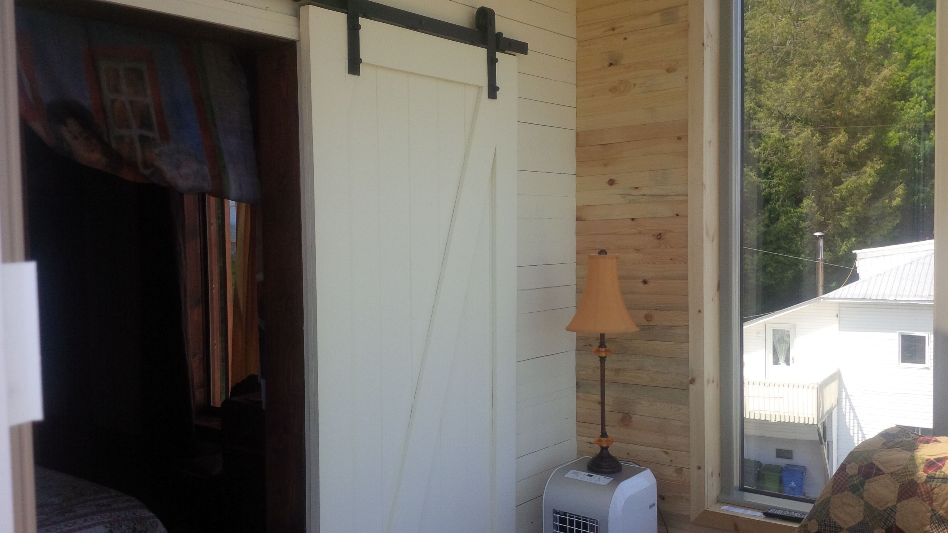 A room with a wooden wall, a white door, a window with a view of trees, and a lamp.