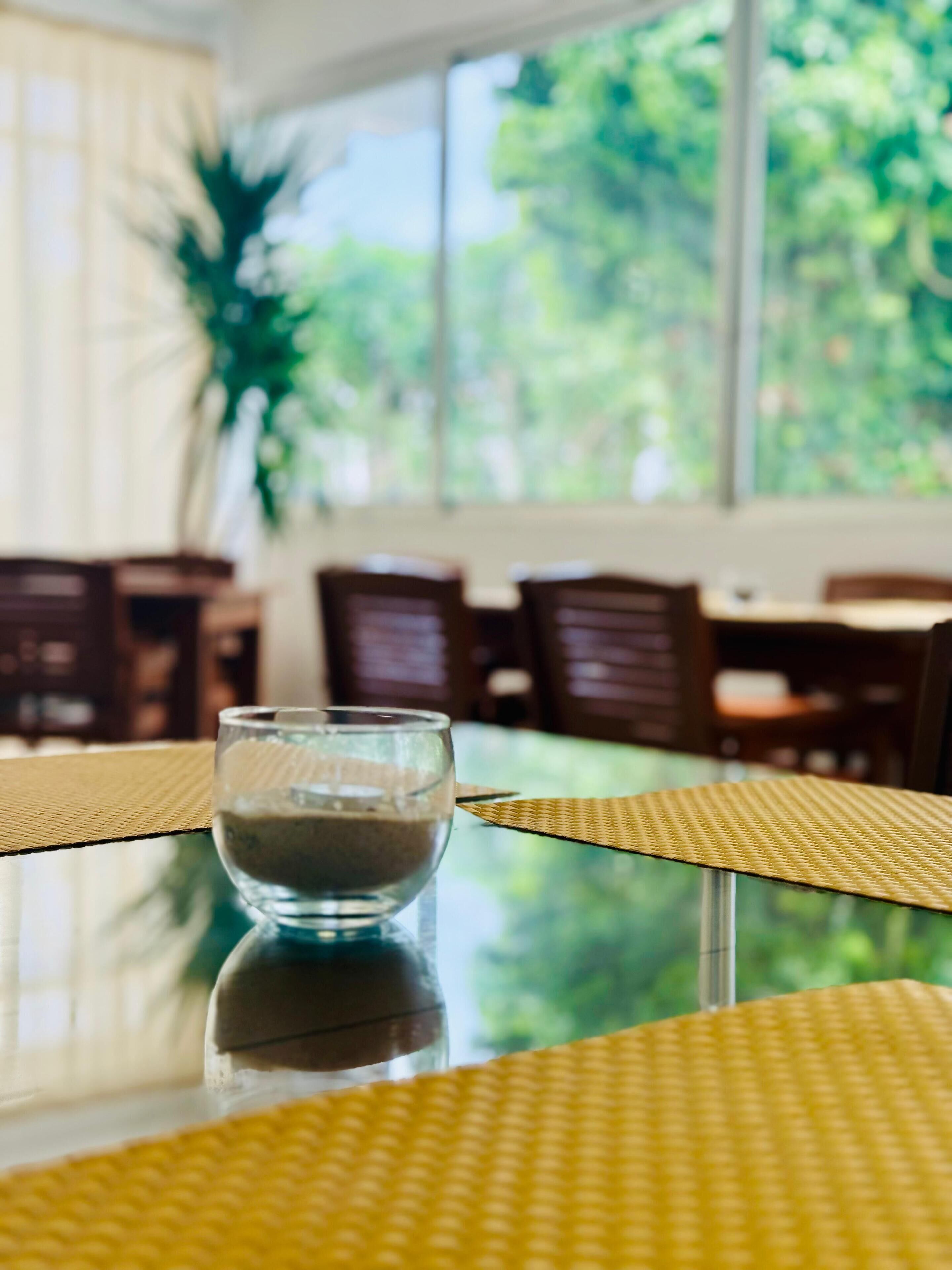 Une table à manger avec un verre d’eau, un set de table en fibres naturelles et une vue sur la verdure grâce à de grandes fenêtres.