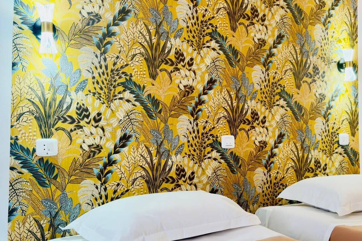 A hotel room with a floral wallpaper, two beds with white and beige bedding, and a wall socket.