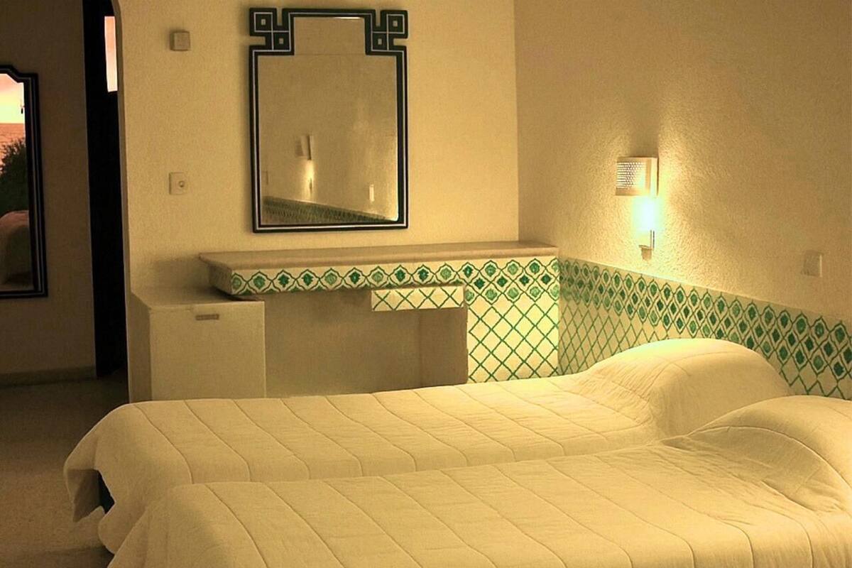 A hotel room with a bed, a nightstand, a mirror, and a wall-mounted light fixture.