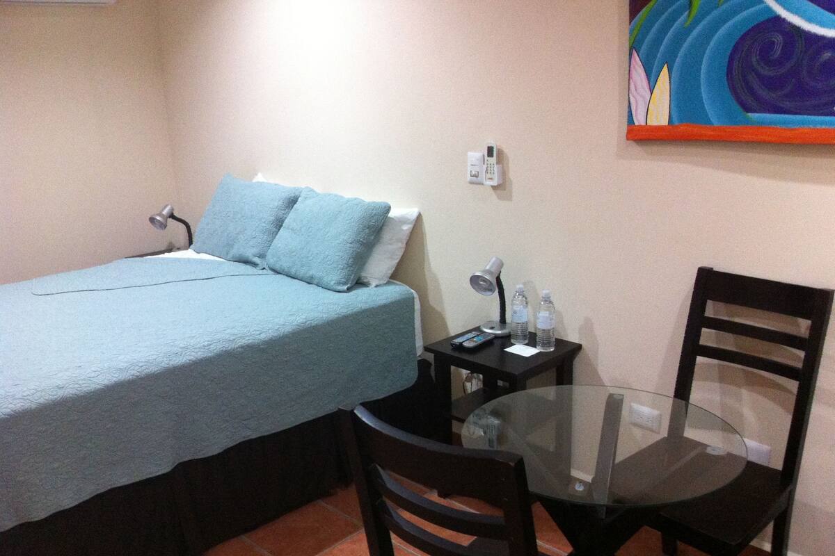 Una habitación pequeña con una cama, una mesa y una silla. Hay una lámpara sobre la mesa y un control remoto a su lado.