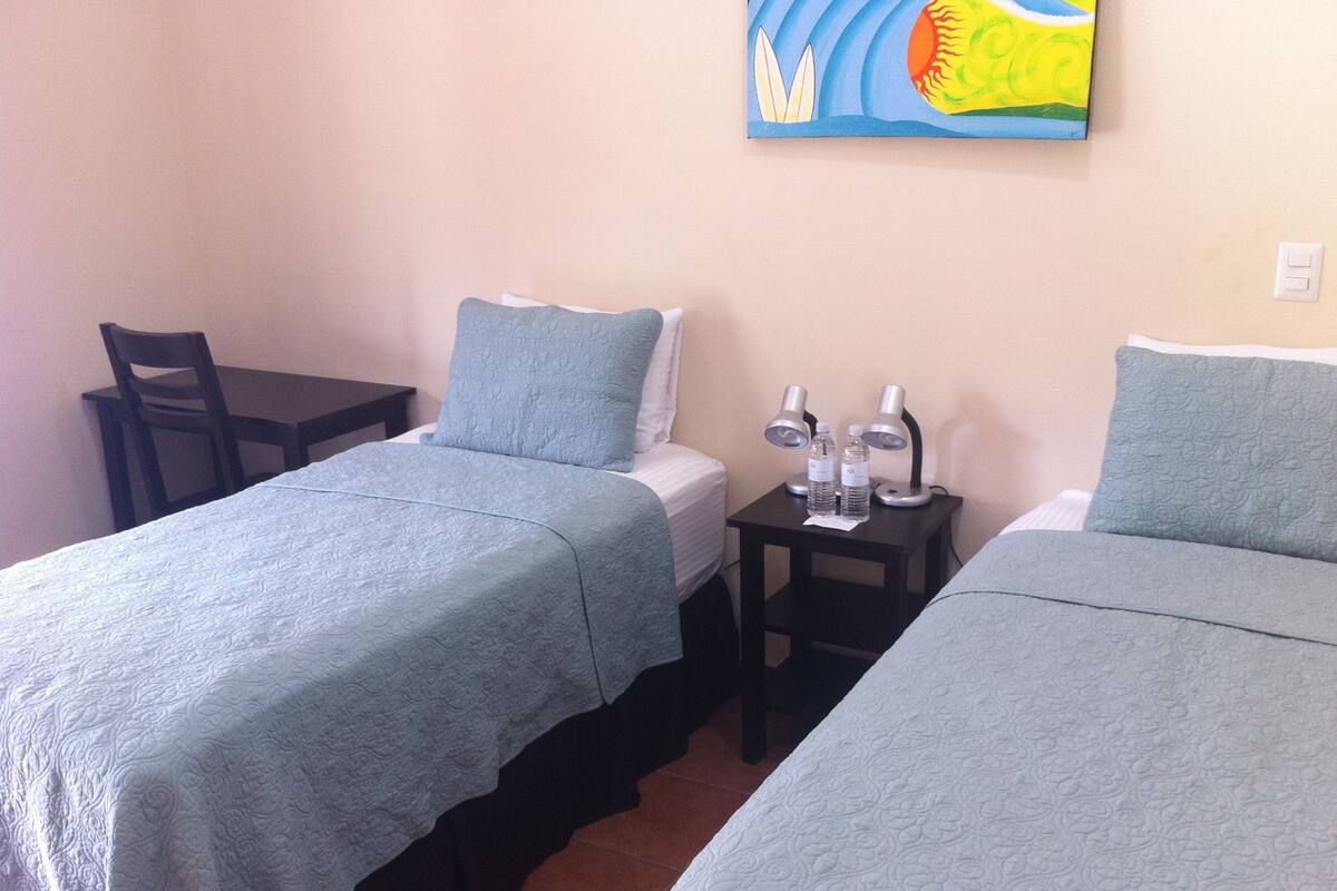 Una habitación con dos camas, una mesita con dos lámparas y una silla.