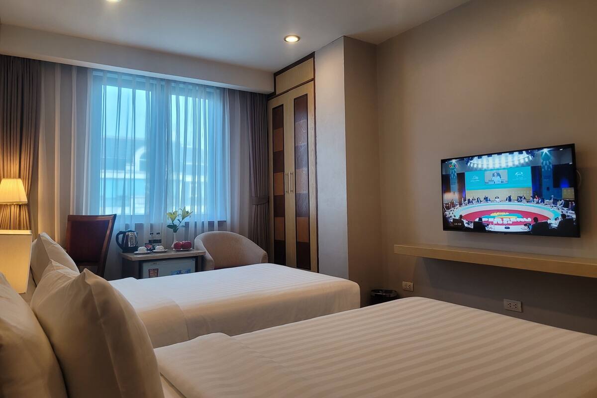 A hotel room with two beds, a TV on the wall, a desk with a lamp, and a window with curtains.