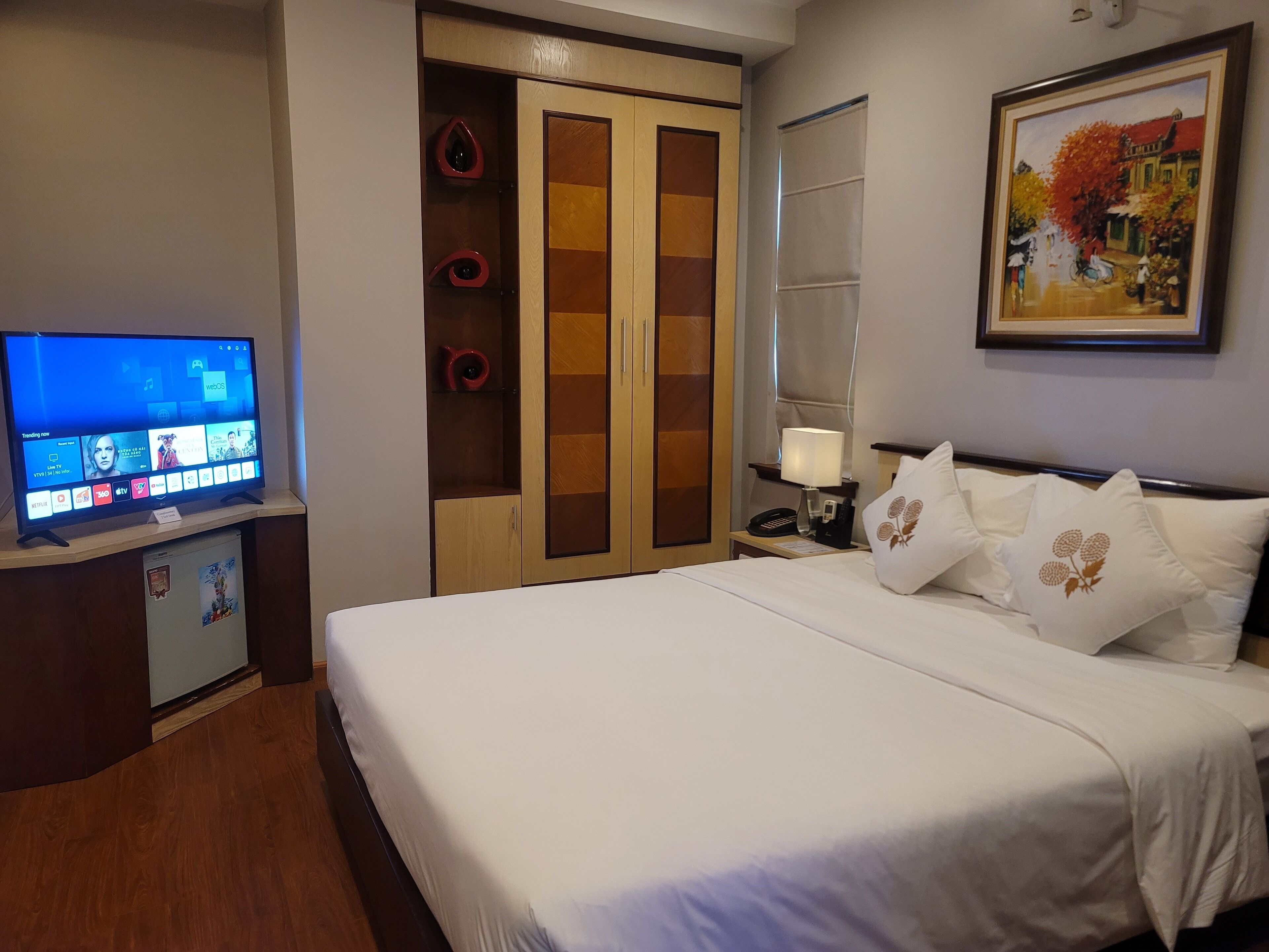 A hotel room with a bed, a TV on a stand, a wardrobe, a nightstand with a lamp, and a framed picture on the wall.