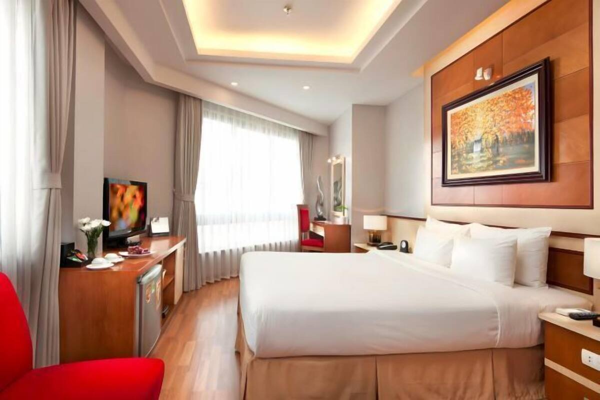 A hotel room with a large bed, a desk with a television, a red chair, and a painting on the wall.