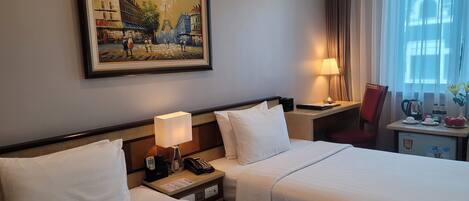A hotel room with two beds, a nightstand with a lamp, a small table with a lamp, a chair, and a painting of a city scene on the wall.