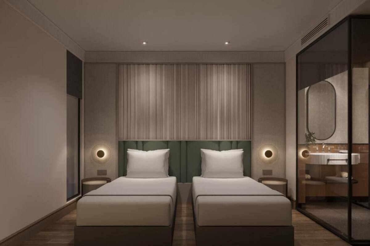 A modern hotel room with two beds, a large mirror, and a glass partition separating the bathroom.