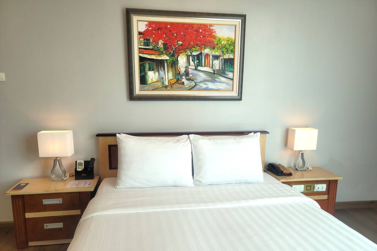 A hotel room with a large bed, two bedside tables with lamps, a painting on the wall, and a wooden nightstand with a phone and a lamp.