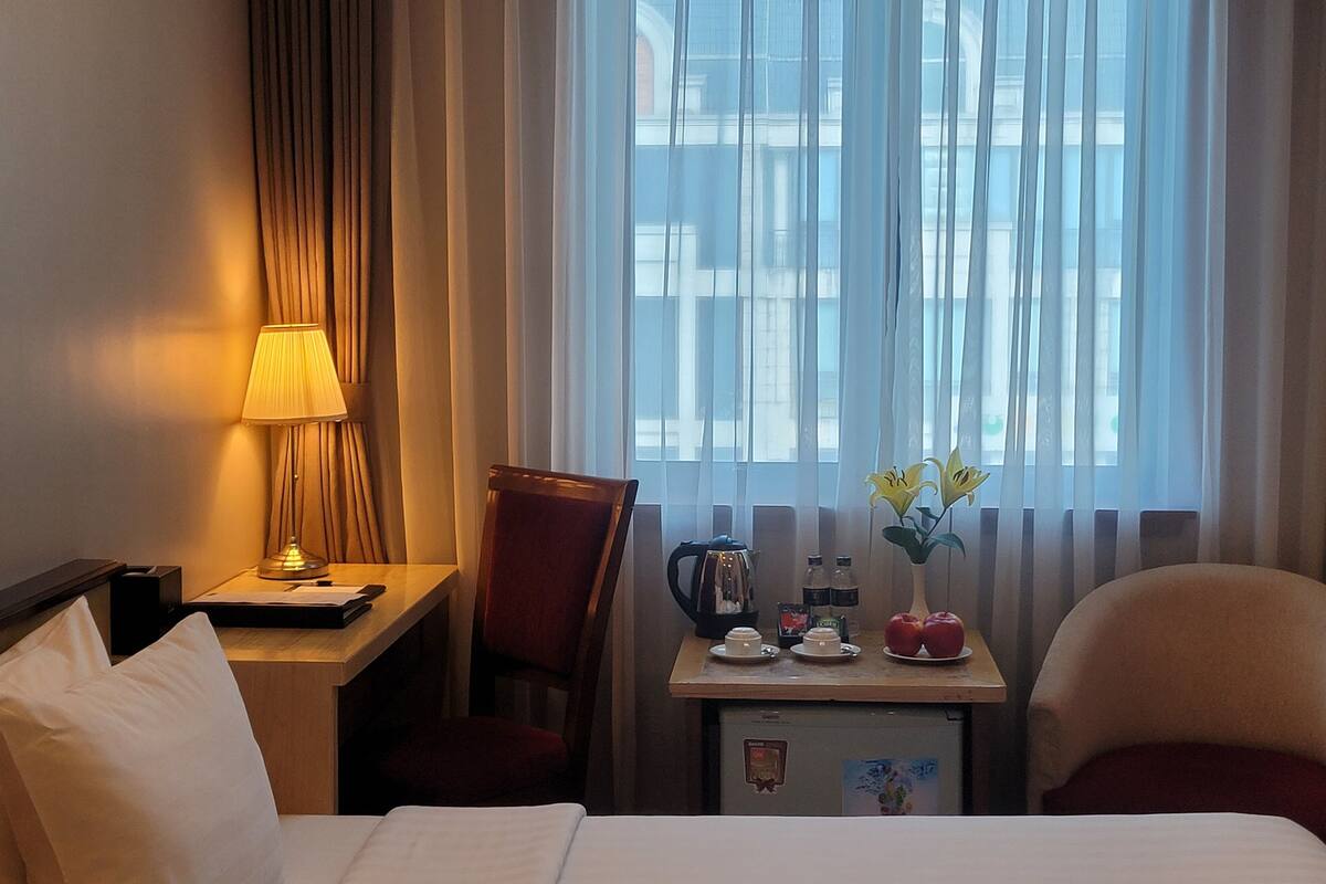 A hotel room with a bed, a desk with a lamp, a chair, a small table with a kettle, a fridge, and a window with curtains.
