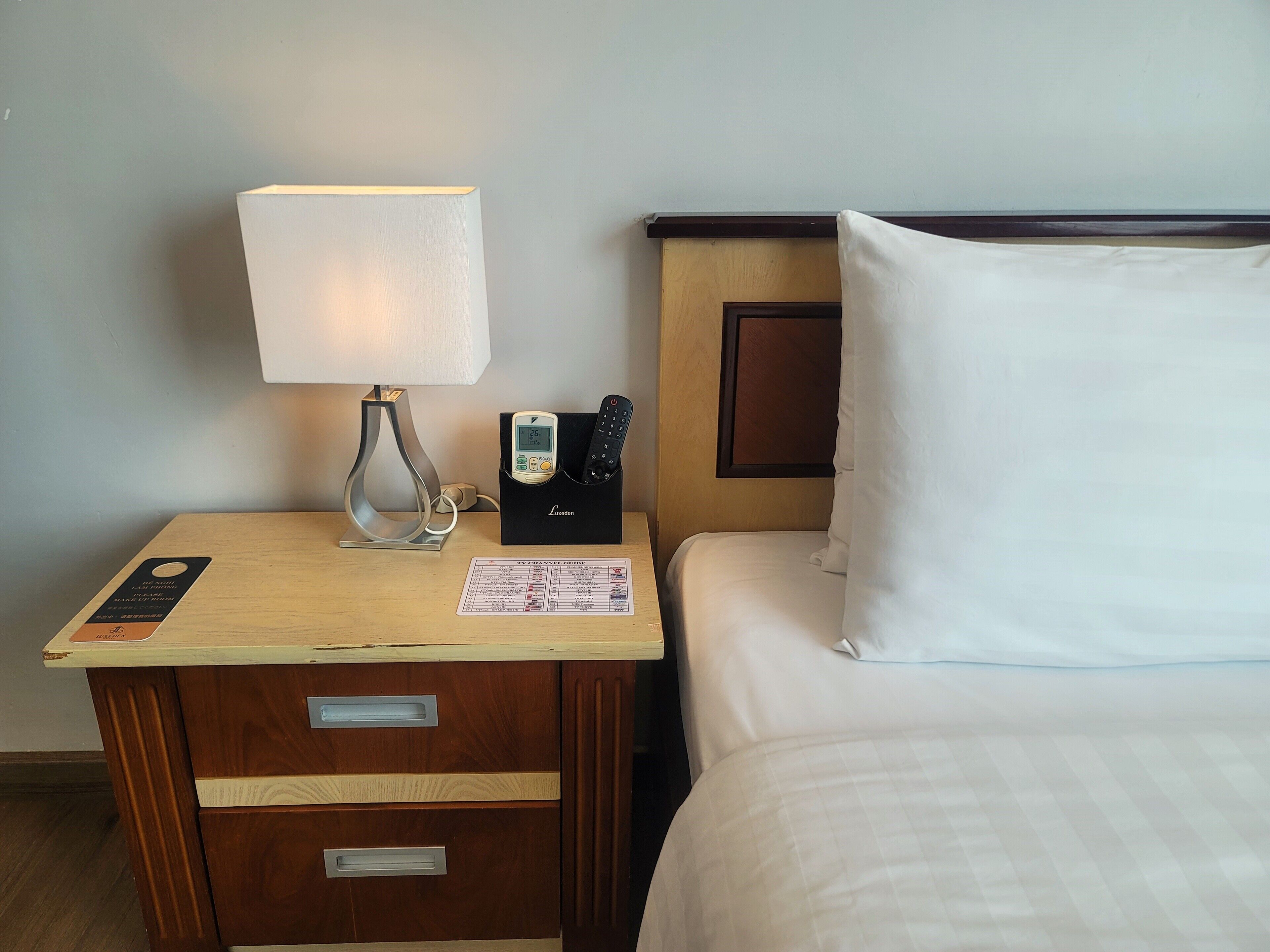 A hotel room with a wooden nightstand, a lamp, a TV channel guide, and a bed with white bedding.