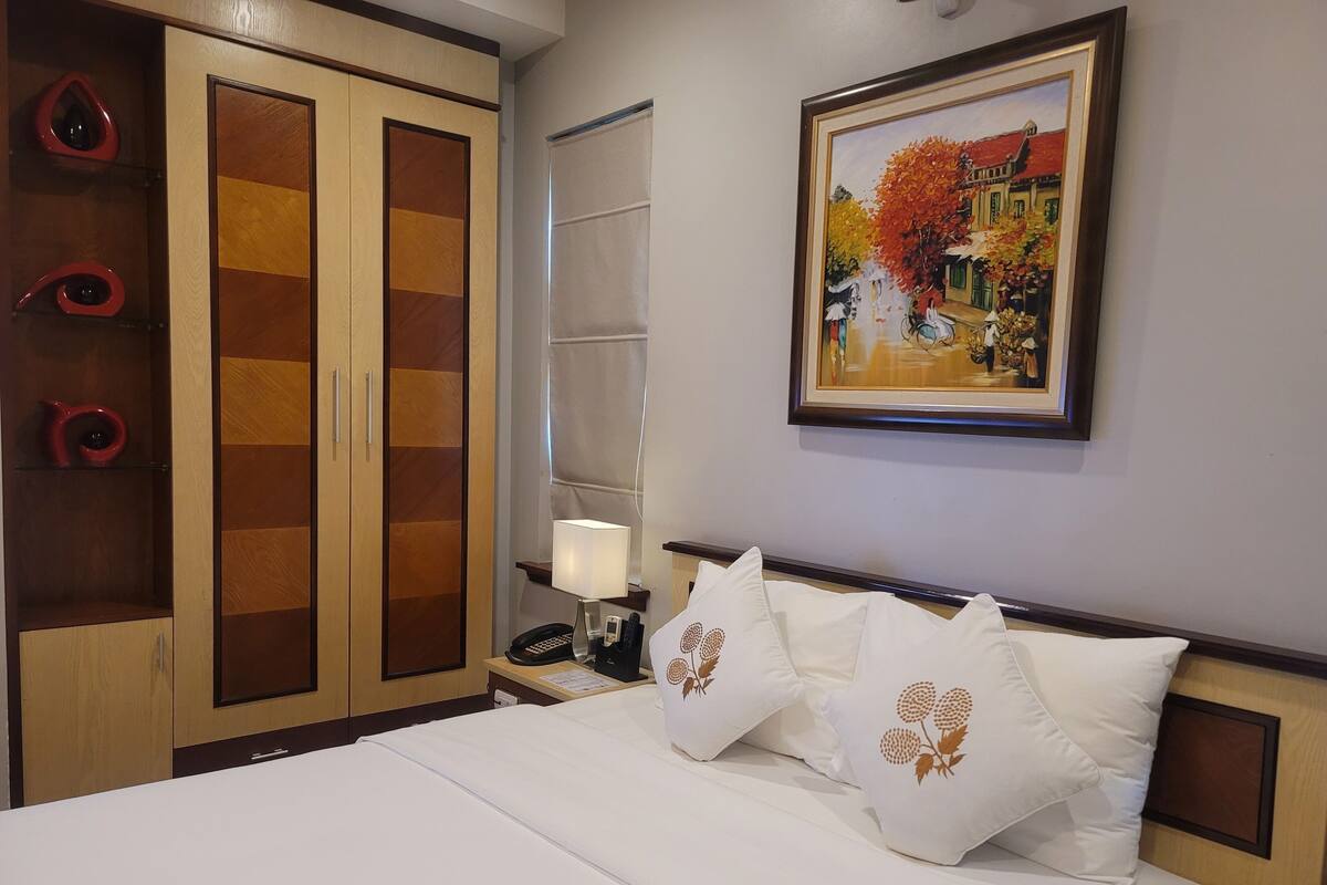 A hotel room with a bed, white bedding, a nightstand with a lamp, a telephone, and a painting on the wall.