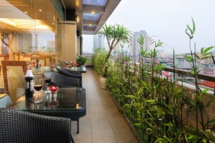 A rooftop terrace with glass tables and chairs, potted plants, and a city view.