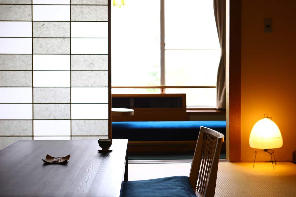 Una habitación con una mesa de madera, un banco tapizado en azul, una lámpara y una ventana con cortinas.