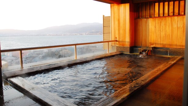 Baño de aguas termales con terraza de madera y vista a un cuerpo de agua y montañas distantes.