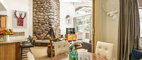 A cozy living room with a stone fireplace, a round wooden table with chairs, and a large window with curtains.