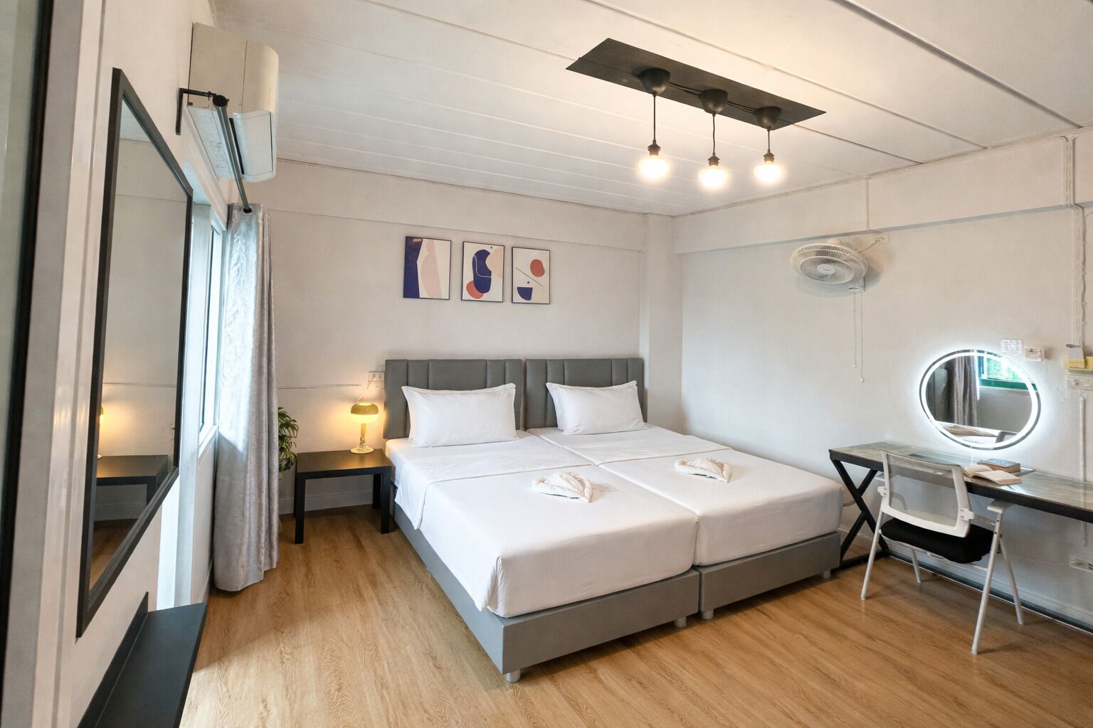A hotel room with a bed, bedside table, lamp, desk with a mirror, and artwork on the wall.