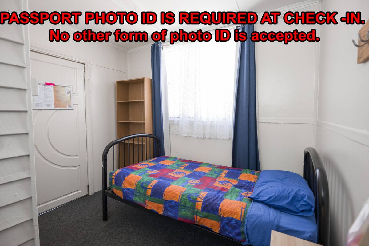 A small, single-bed room with a colorful quilt, a chair, a wooden shelf, and a door with a notice.
