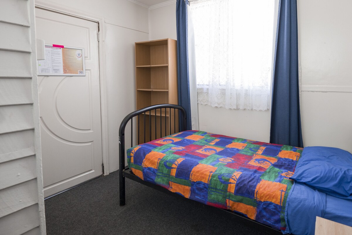 Una habitación pequeña con cama individual, colcha colorida, una silla, estante de madera y puerta con un aviso.