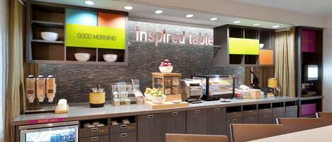 Una barra de desayuno con un cartel que dice "inspired table", tazones, un refrigerador con bebidas y una máquina de café.