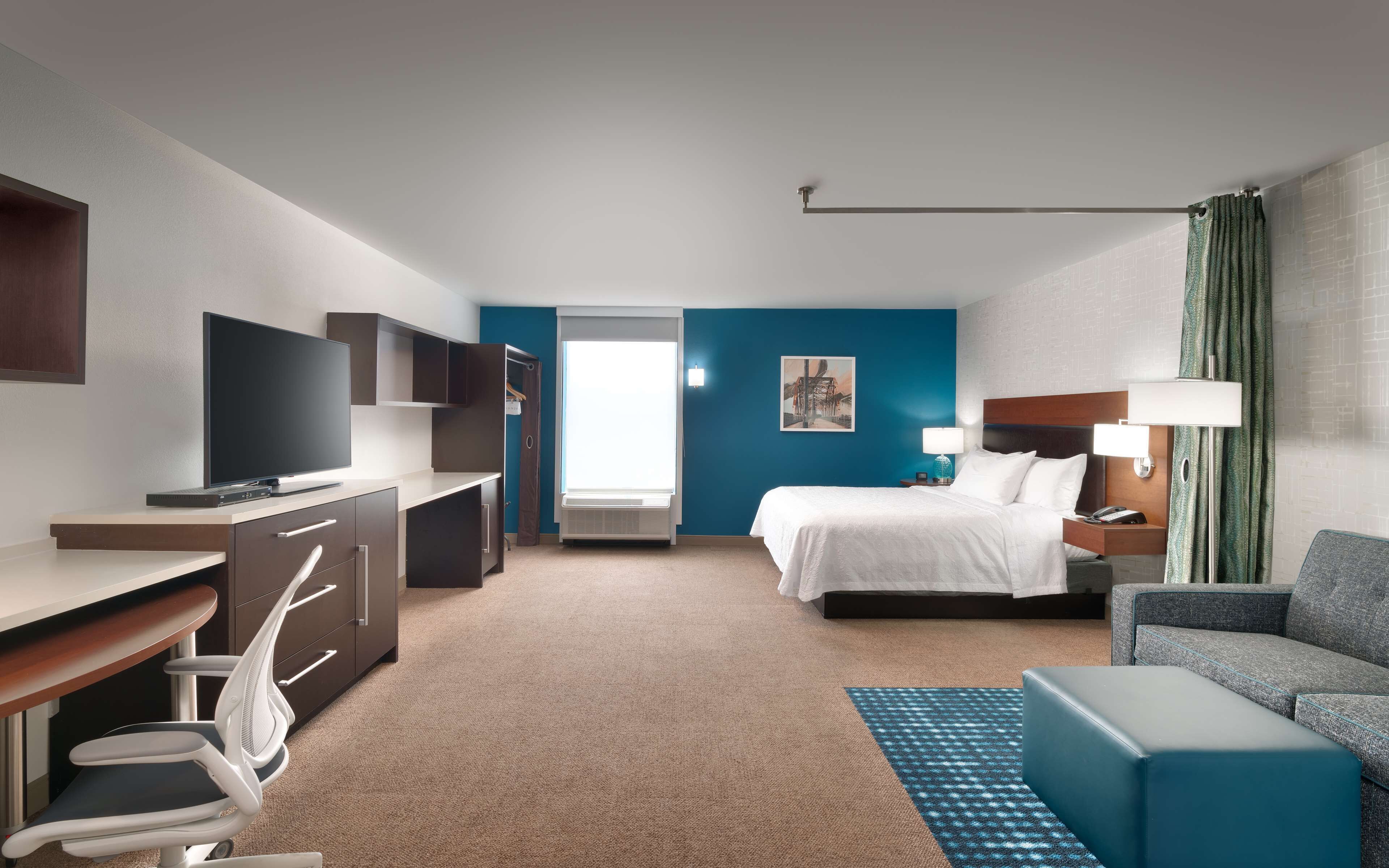 A hotel room with a bed, a desk with a TV, a chair, a sofa, and a window with curtains.