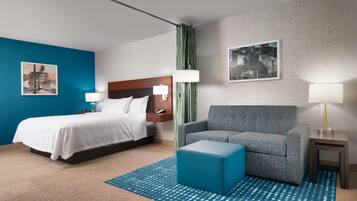 Une chambre d’hôtel avec un lit, un canapé gris, un pouf bleu, une petite table et deux lampes fixées au mur.