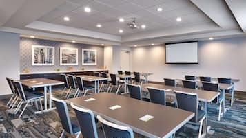 A conference room with multiple tables and chairs, a projector screen, and framed pictures on the wall.