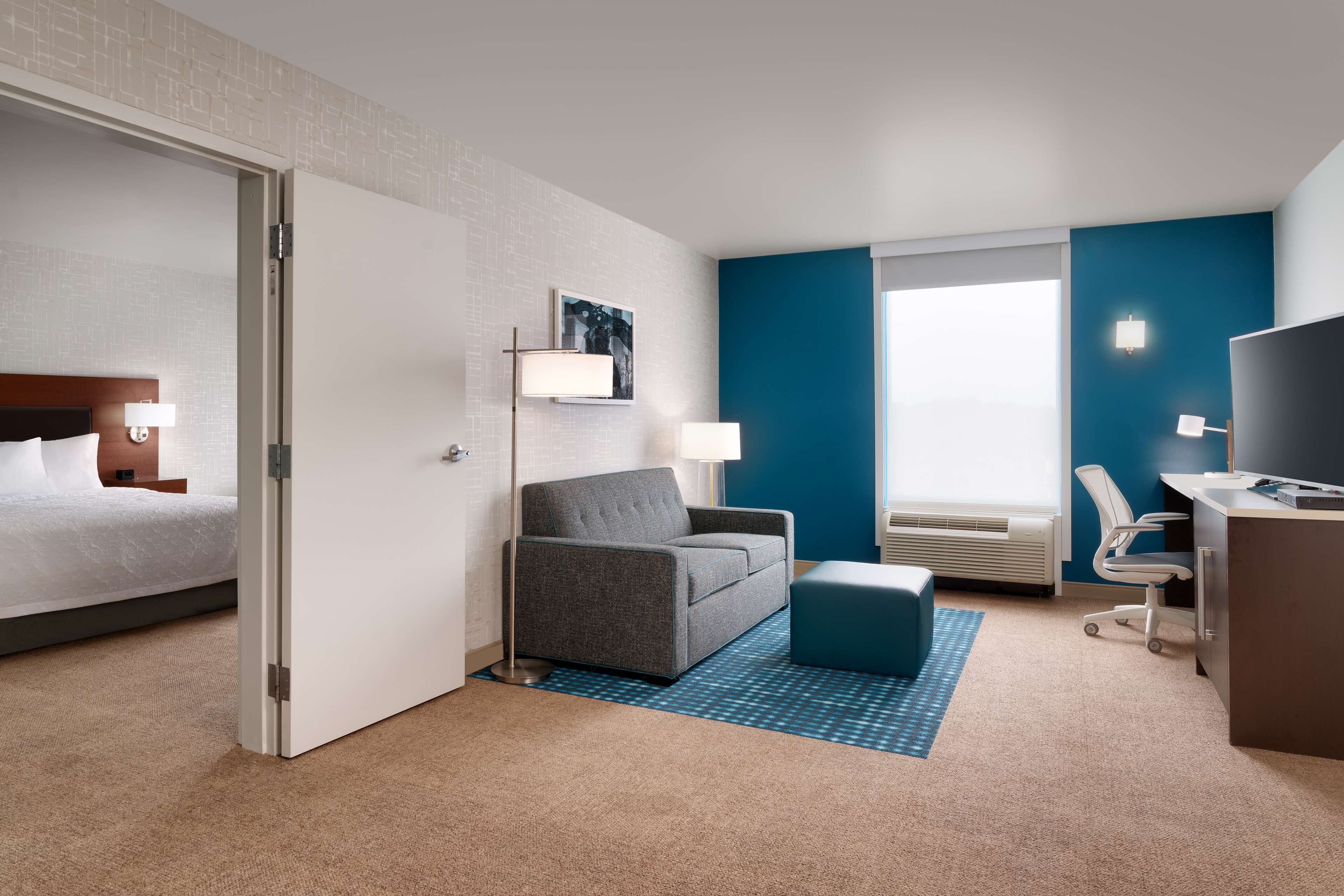 Una habitación de hotel con una cama, un sofá gris, un escritorio con una silla, un televisor y una ventana con vistas.