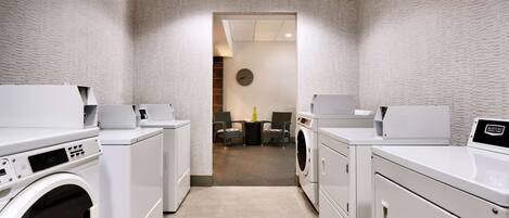 A laundry room with white appliances, a clock on the wall, and a view into another room.