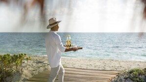 A man in a white shirt and hat carrying a tray with bottles and a glass on a wooden boardwalk by the beach.