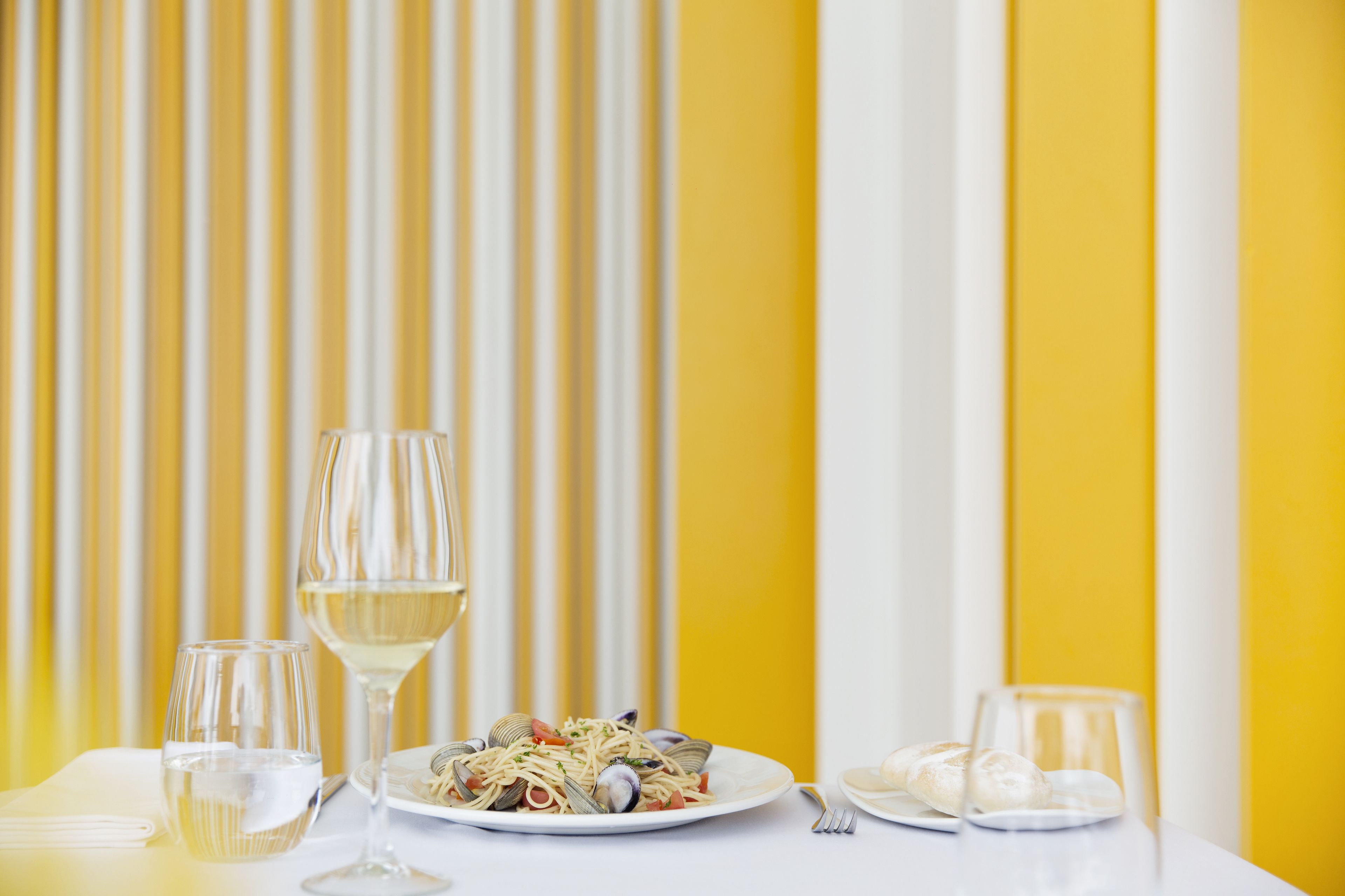 A dining table with a plate of pasta, two glasses of white wine, and a fork.