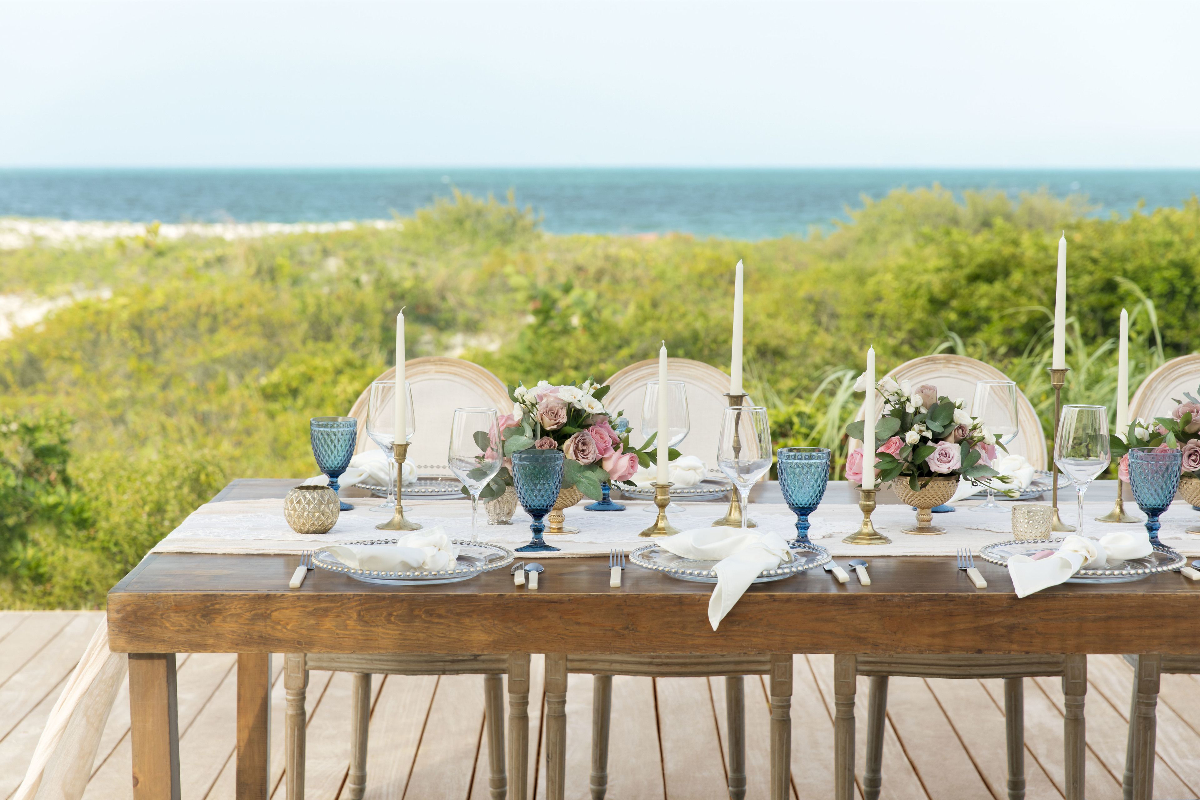 Ein Holztisch, gedeckt für eine Mahlzeit, mit blauem Glas, weißen Servietten und Kerzen, mit Blick auf einen Strand.