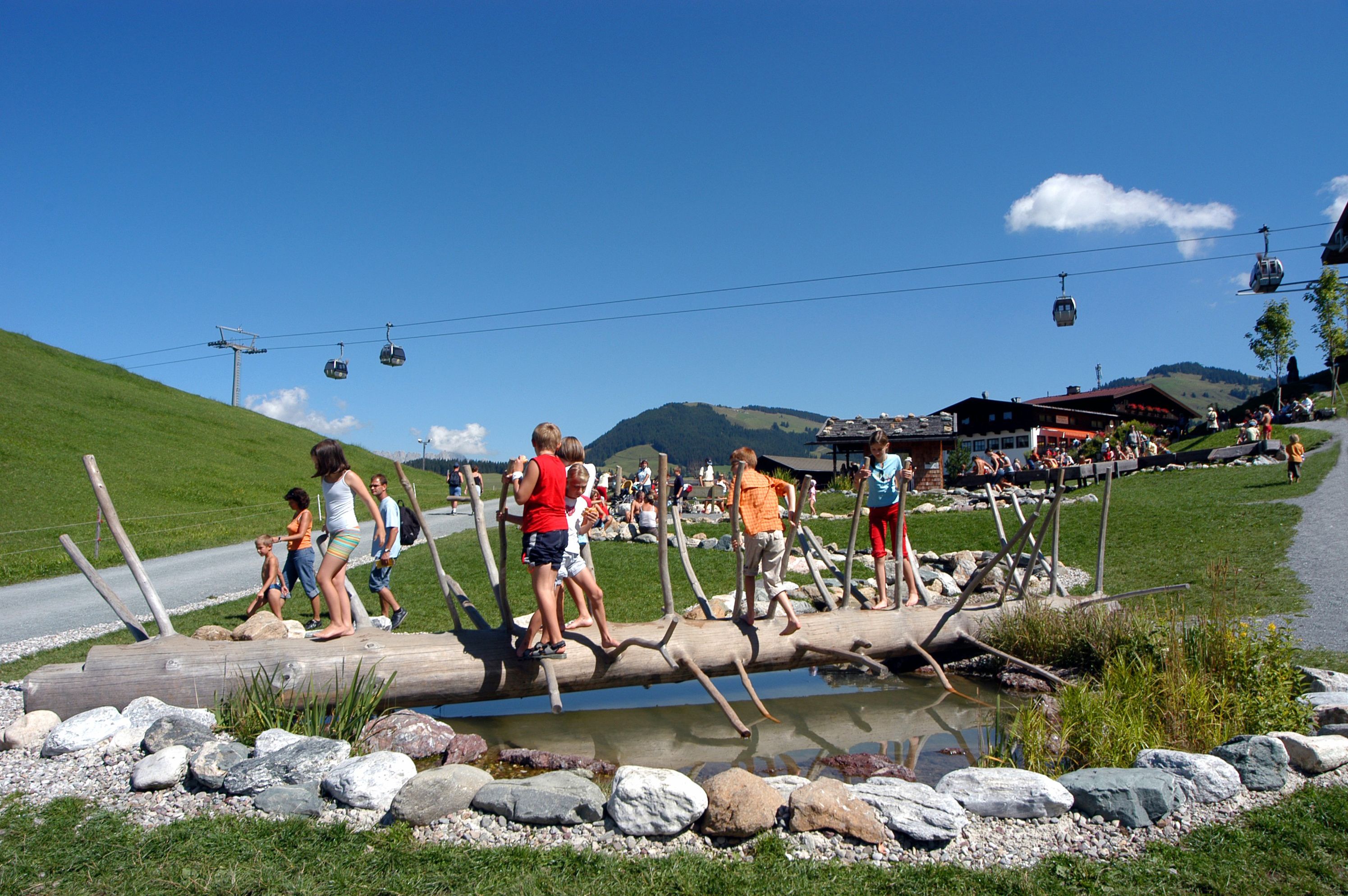 Un ponte di tronchi sopra un ruscello con persone che lo attraversano, una seggiovia sullo sfondo e una montagna all'orizzonte.
