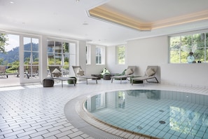 A modern indoor swimming pool with a tiled floor, surrounded by lounge chairs and a view of a mountainous landscape through large windows.