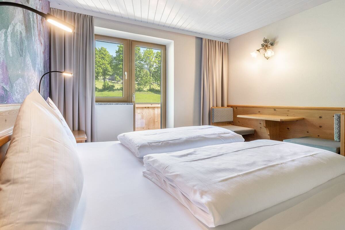 A hotel room with two beds, a wooden headboard, a window with curtains, and a wall-mounted light fixture.