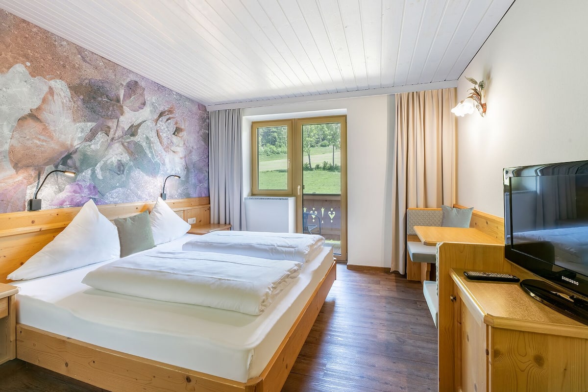 A hotel room with a bed, a desk with a TV, a window with curtains, and a mural on the wall.
