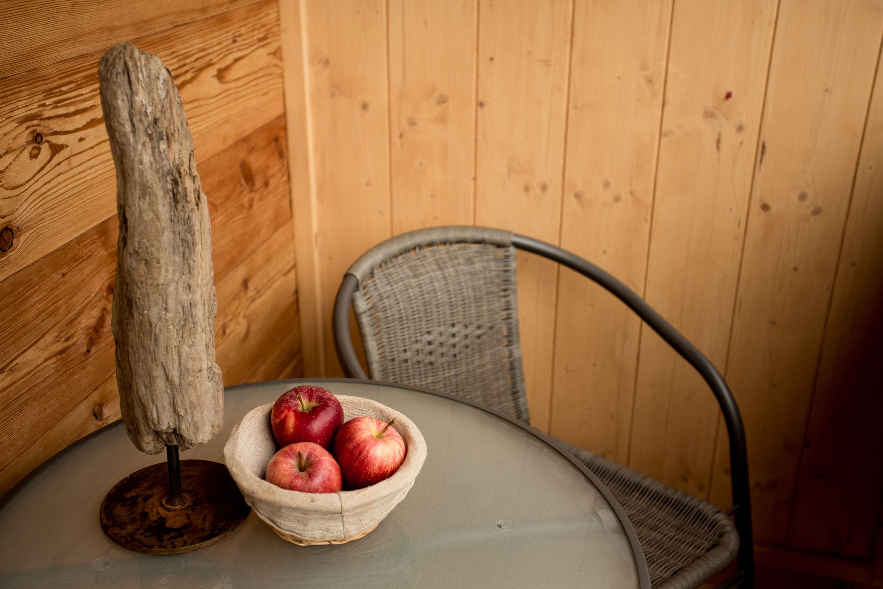 A table with a woven basket containing apples, a rustic wooden object, and a wicker chair.