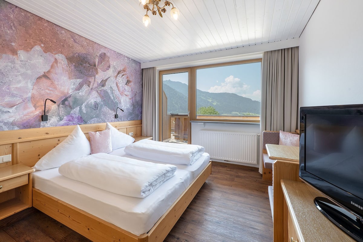 A hotel room with two beds, a large window with a mountain view, a television, and a wall with a floral mural.